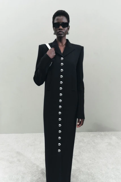 An African American woman poses in a long black coat with silver buttons, wearing sunglasses and carrying a white purse indoors.
