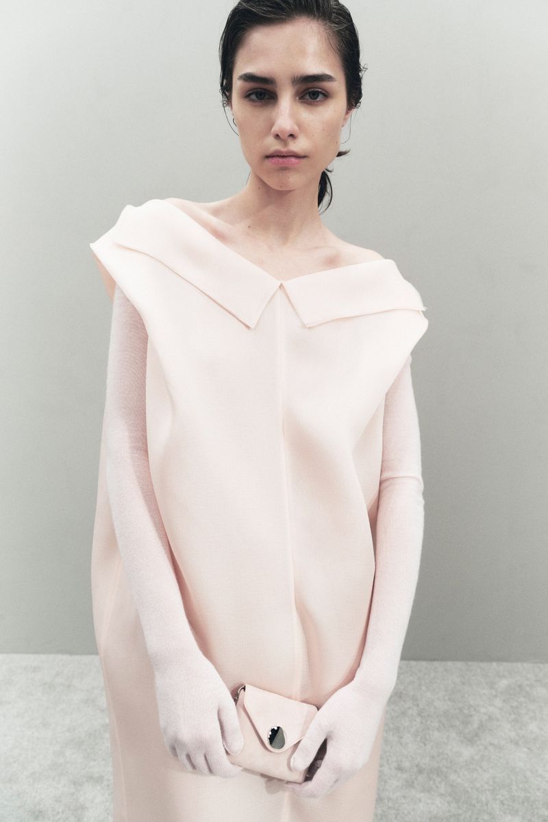 Fashion shot of a female model in a light pink dress and matching purse, posing in an indoor studio with a neutral background.