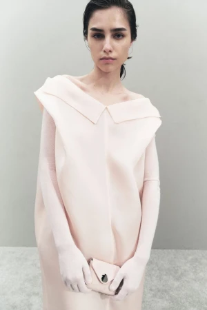 Fashion shot of a female model in a light pink dress and matching purse, posing in an indoor studio with a neutral background.