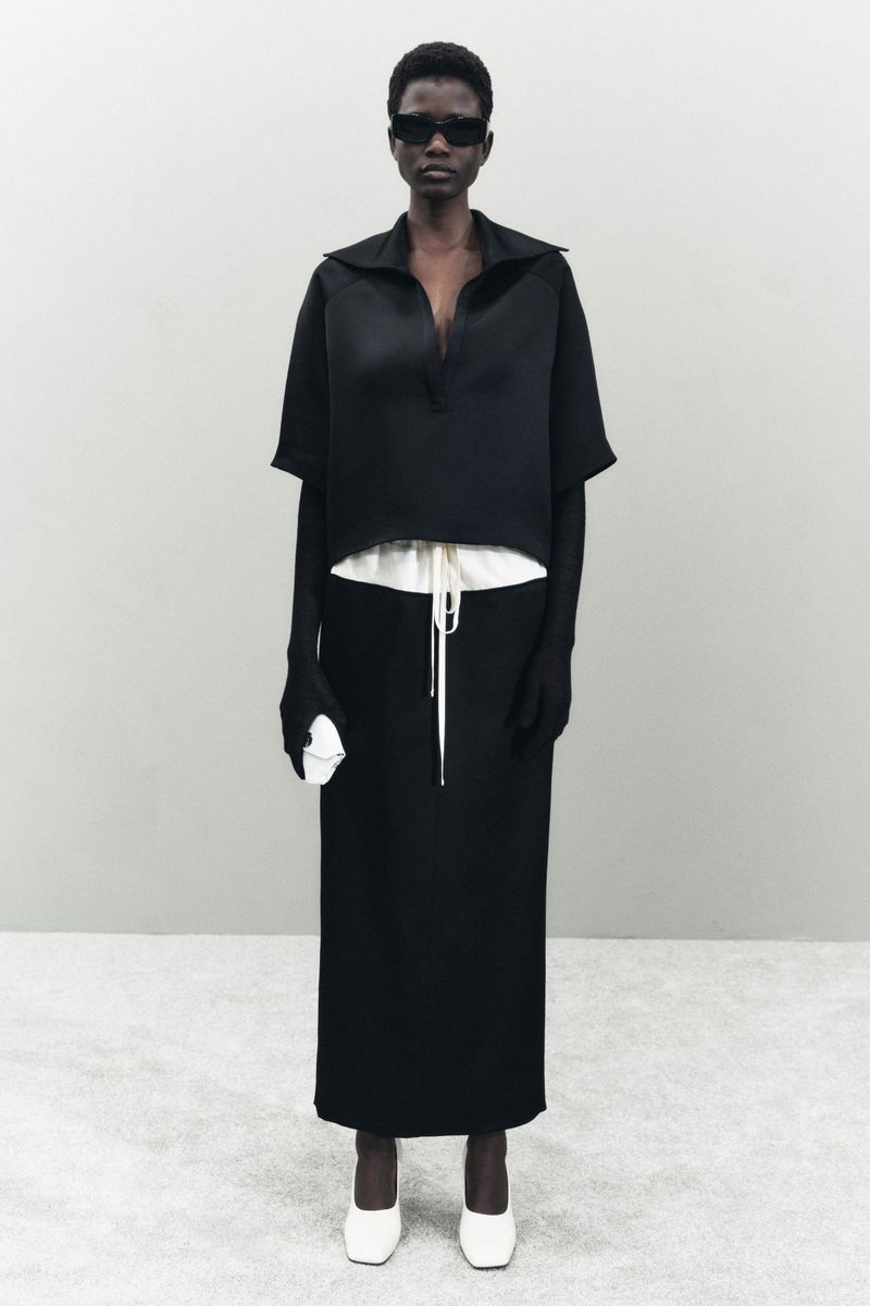 Dark-skinned fashion model in black top and maxi skirt with white shoes, wearing sunglasses. Minimalist fashion studio shot.