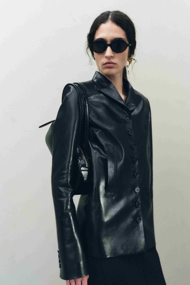 Adult female wearing a black leather blazer, black skirt, and black sunglasses, posing indoors against a plain off-white wall.