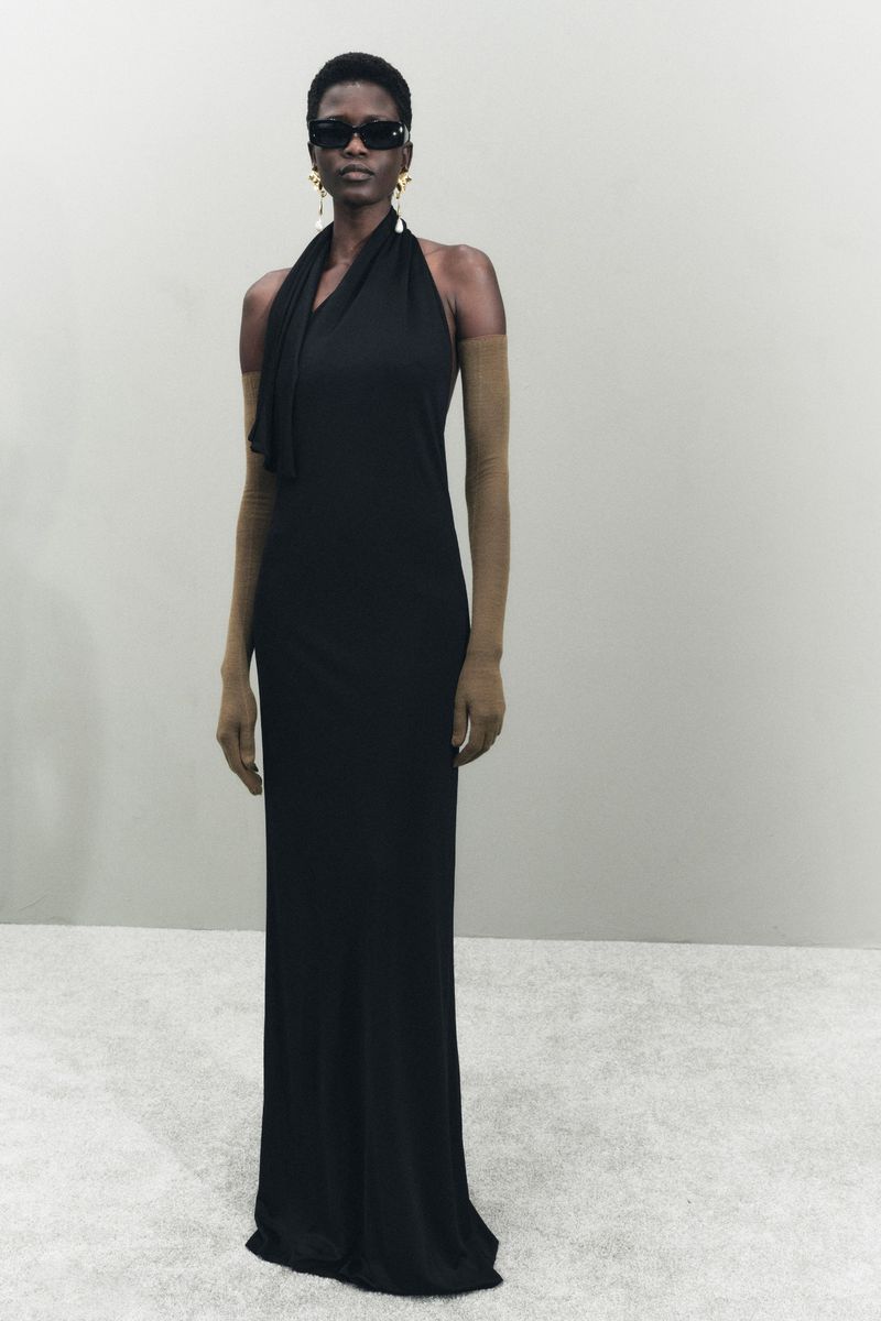 Full-length portrait of a woman in a black halter dress, sunglasses, and gold earrings, standing in a neutral studio setting.