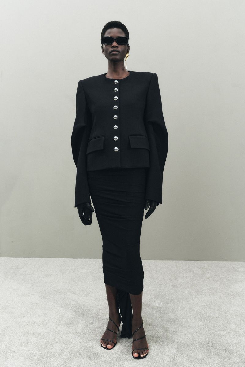 A slender Black woman posing in a sleek black jacket, matching skirt, strappy heels, and sunglasses against a neutral background.