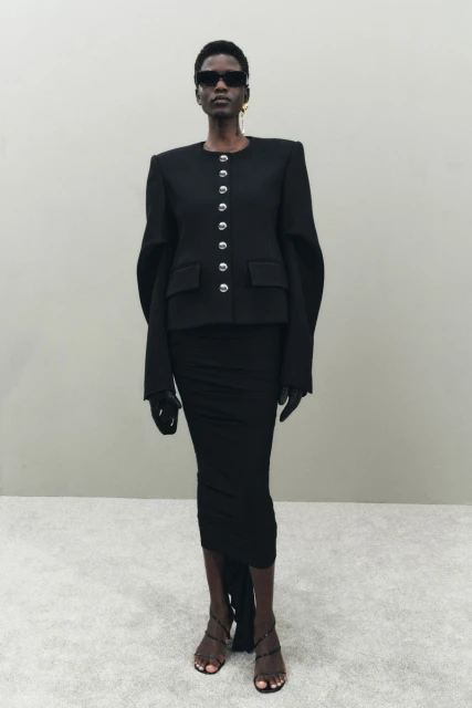 A slender Black woman posing in a sleek black jacket, matching skirt, strappy heels, and sunglasses against a neutral background.