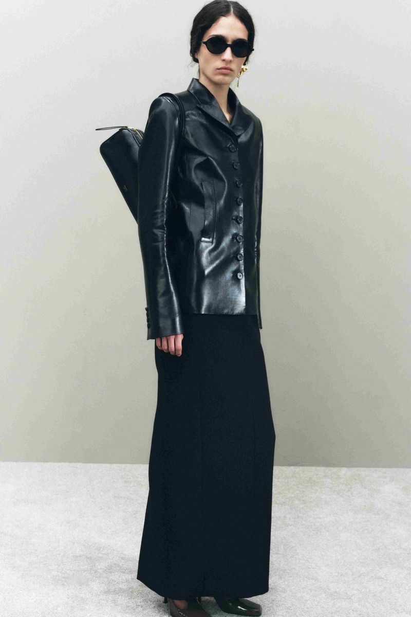 Fashionable woman in sunglasses poses in leather jacket and maxi skirt, holding a black bag against a neutral backdrop.