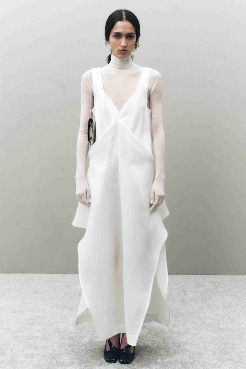 Elegant fashion shot of a woman in a white dress with sheer gloves against a neutral background, showcasing contemporary style.