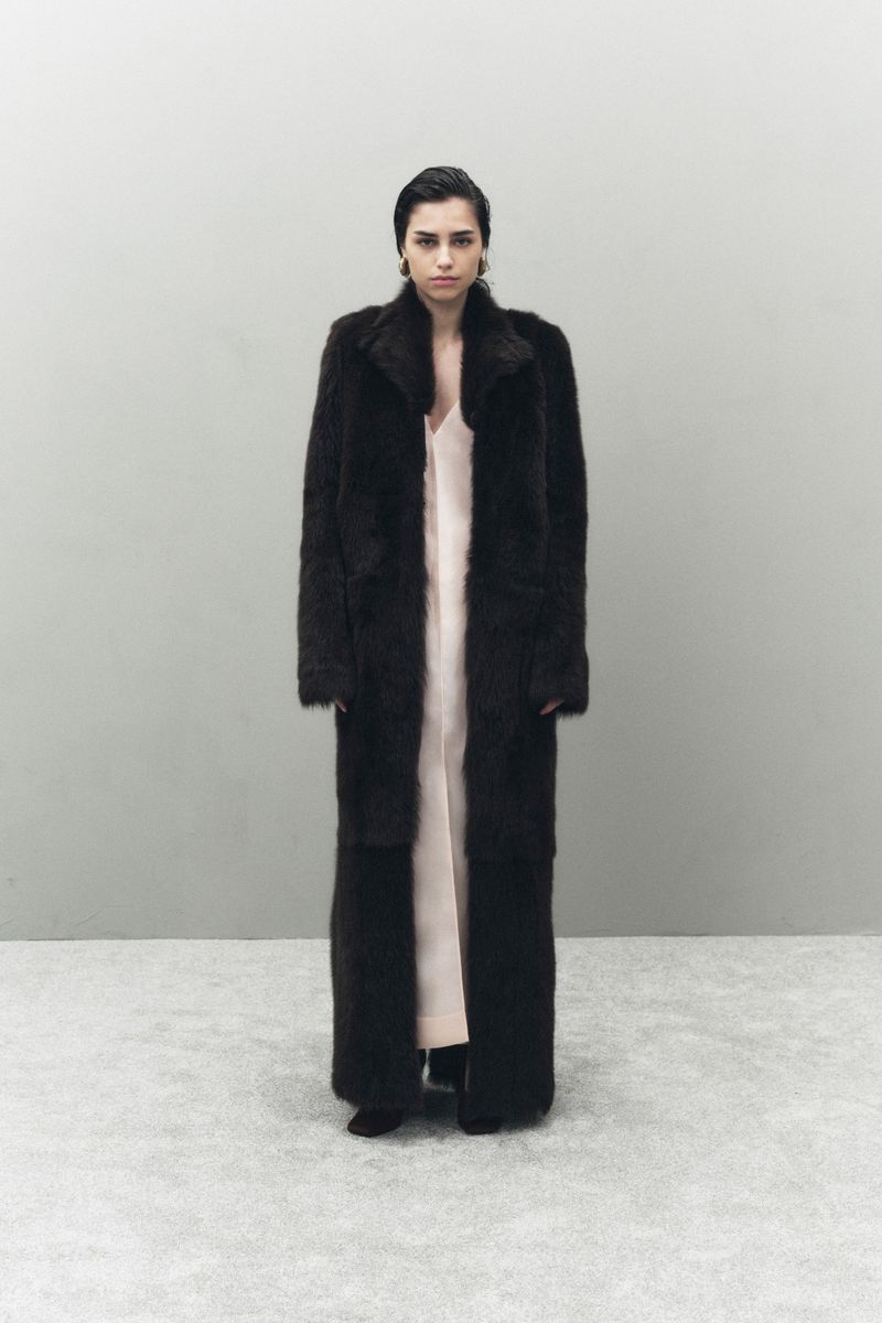 A woman in a full-length dark coat and light dress stands in front of a grey wall. The portrait is well-lit, with minimal shadows.