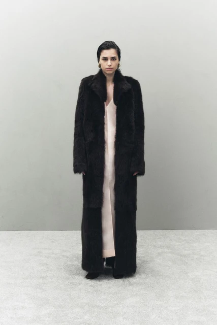 A woman in a full-length dark coat and light dress stands in front of a grey wall. The portrait is well-lit, with minimal shadows.