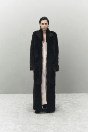 A woman in a full-length dark coat and light dress stands in front of a grey wall. The portrait is well-lit, with minimal shadows.