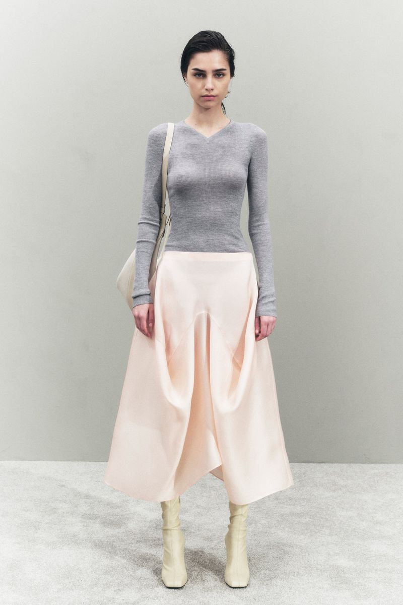 A young woman poses in a studio wearing a gray sweater, pale pink skirt, and off-white boots, carrying an off-white shoulder bag.