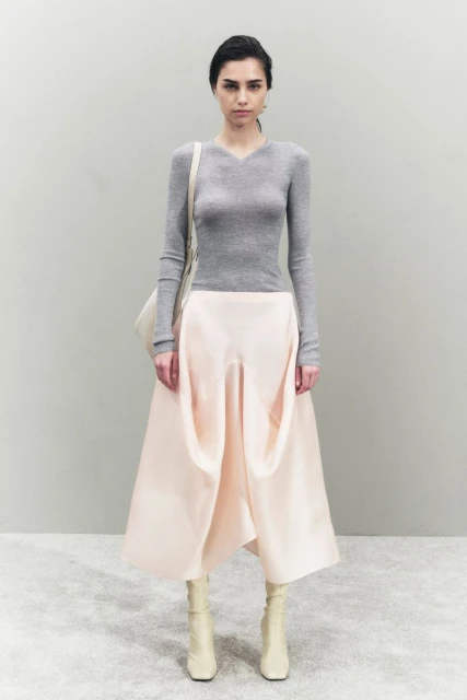 A young woman poses in a studio wearing a gray sweater, pale pink skirt, and off-white boots, carrying an off-white shoulder bag.