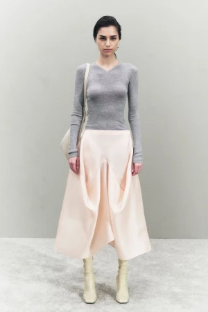 A young woman poses in a studio wearing a gray sweater, pale pink skirt, and off-white boots, carrying an off-white shoulder bag.