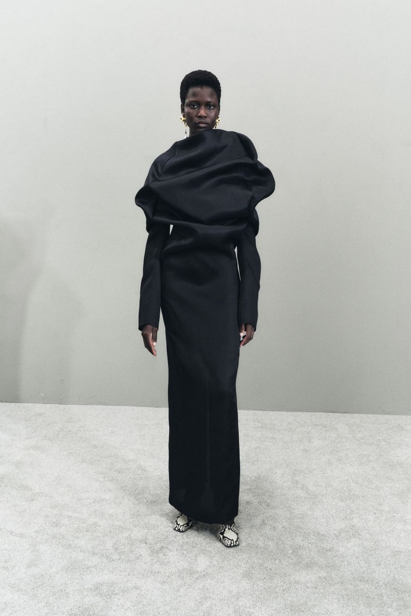 An adult Black female fashion model wears a sculptural black gown, accessorized with gold earrings, standing in front of a light grey wall.