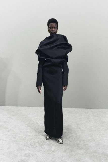 An adult Black female fashion model wears a sculptural black gown, accessorized with gold earrings, standing in front of a light grey wall.