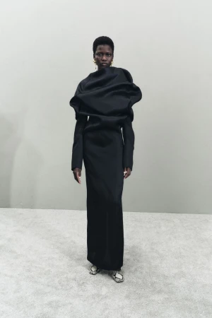 An adult Black female fashion model wears a sculptural black gown, accessorized with gold earrings, standing in front of a light grey wall.