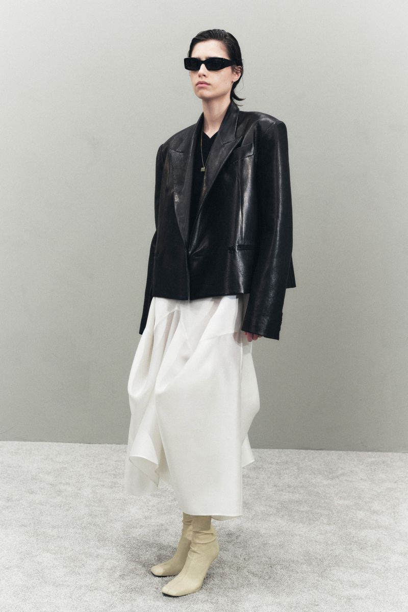Fashionable woman in a black leather blazer, white skirt, sunglasses, and heeled boots, posing in a photo studio with a neutral backdrop.