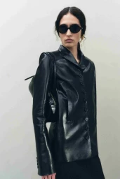 Model in dark sunglasses and black leather coat carrying a shoulder bag against a white seamless background