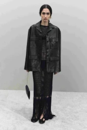 Model wearing a black leather utility jacket and mesh dress holding a small clutch against a grey seamless studio background.