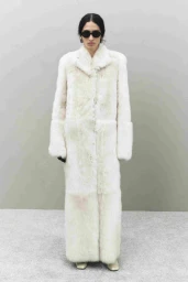 Model wearing a long white textured coat and dark sunglasses against a seamless grey backdrop.