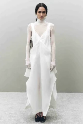 Model wearing a white dress over a sheer turtleneck against a neutral grey wall and floor studio background