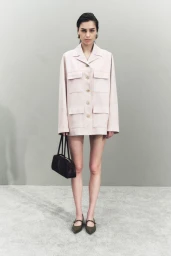 Model wearing a light pink utility jacket and carrying a dark handbag against a plain grey studio backdrop