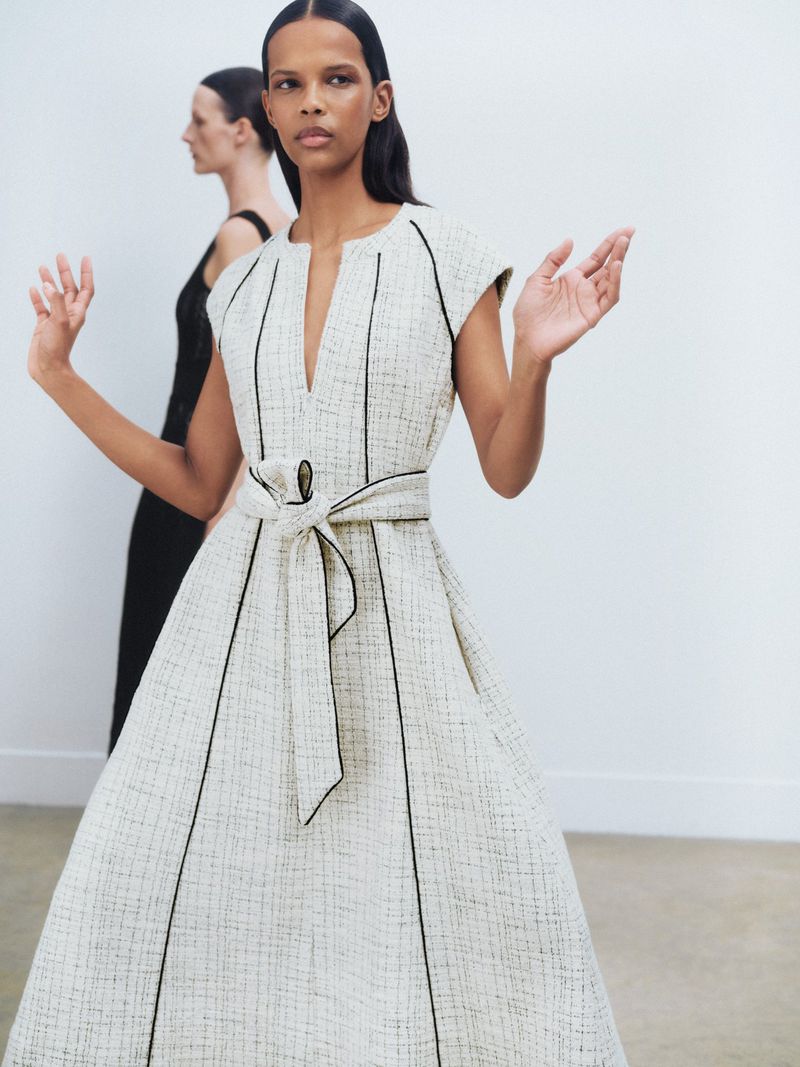 Two women pose indoors in a fashion shot. One stands in front wearing a cream-colored dress with black trim and a tied waist.