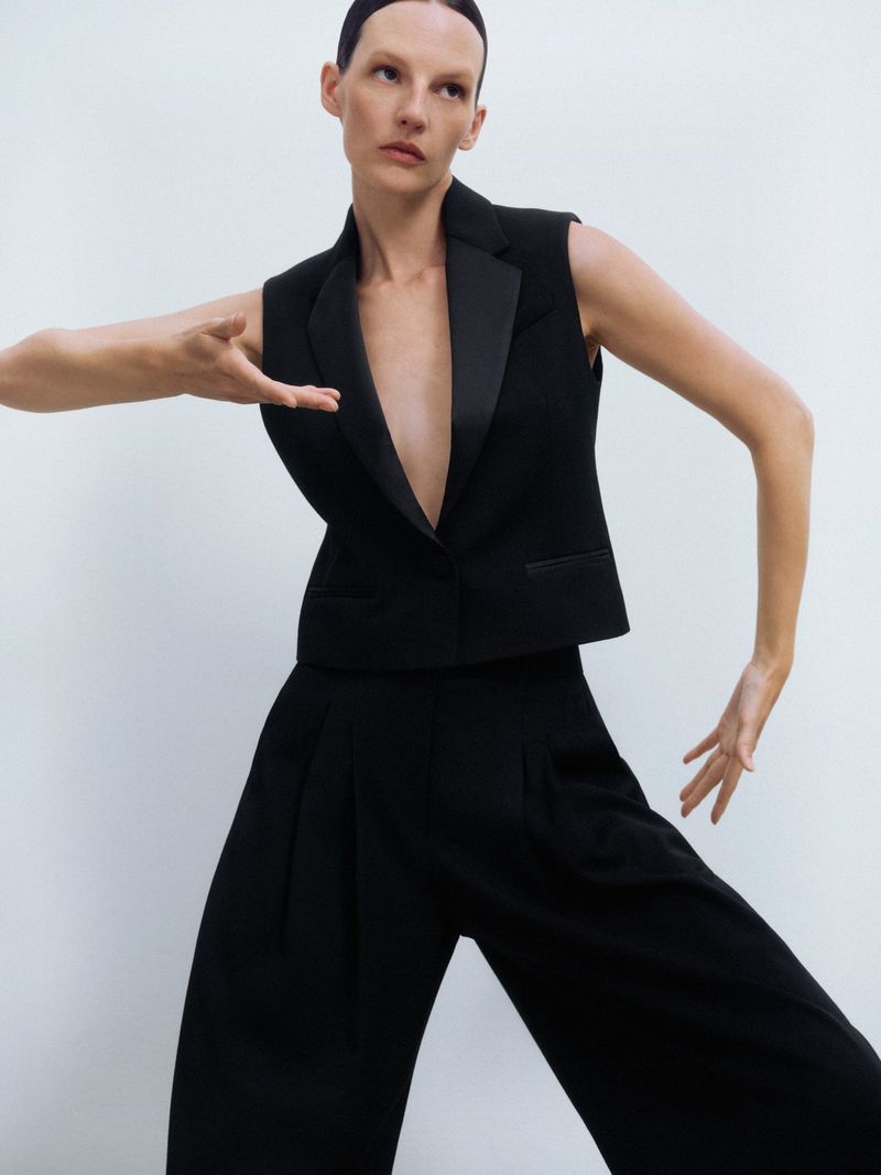 A fashion photo showcasing a poised adult female model in a black vest and culottes, against a bright white backdrop. Emphasizing minimalist styling.