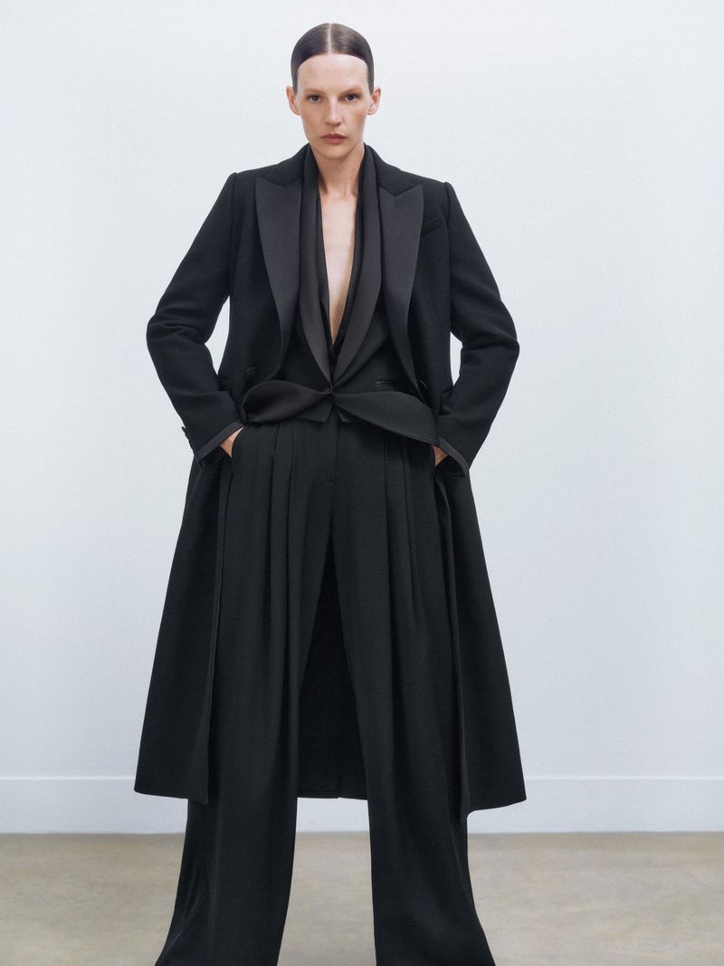 A woman in a black blazer, vest, and wide-leg pants poses in front of a white wall. The outfit has a sleek, modern, and sophisticated design.