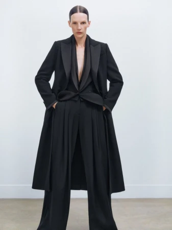 A woman in a black blazer, vest, and wide-leg pants poses in front of a white wall. The outfit has a sleek, modern, and sophisticated design.
