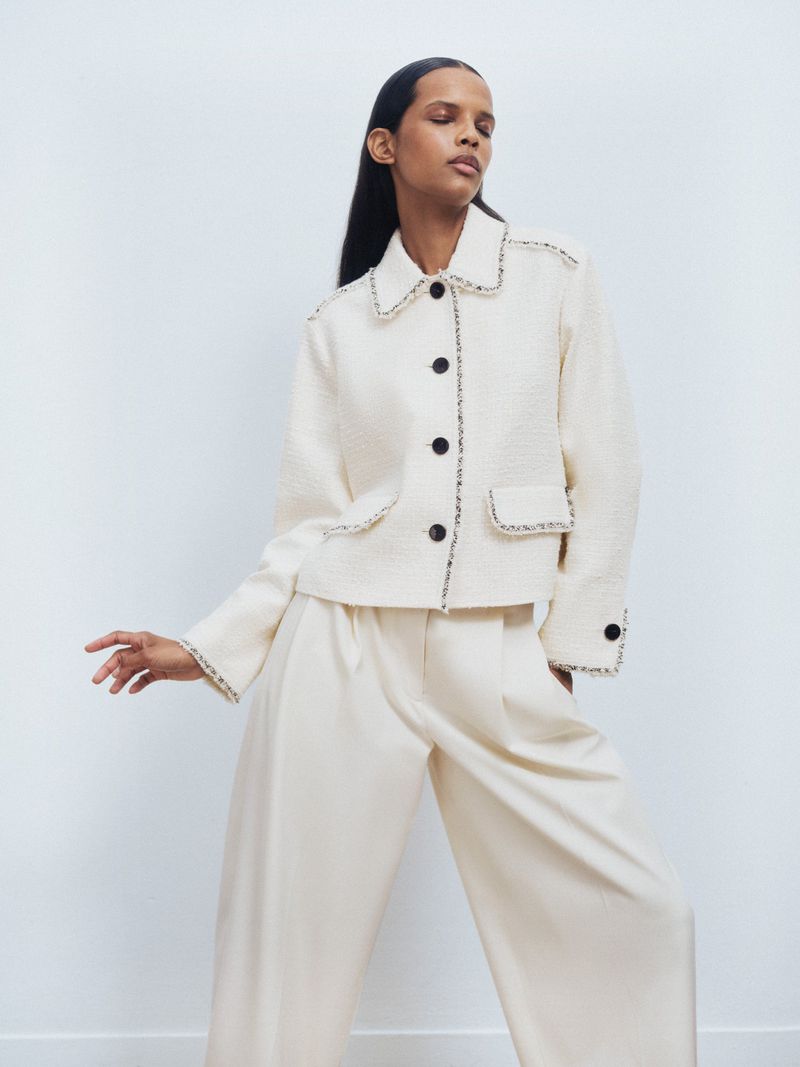 Adult fashion model poses in an off-white suit jacket and matching wide-leg trousers against a bright white studio backdrop.
