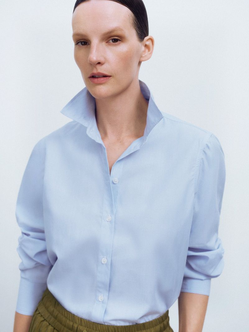 A woman with dark hair pulled back is wearing a light blue collared shirt and olive green pants against a white backdrop in a studio shot.