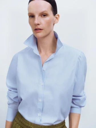 A woman with dark hair pulled back is wearing a light blue collared shirt and olive green pants against a white backdrop in a studio shot.