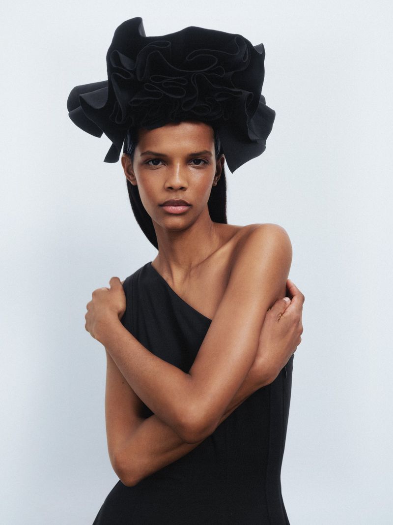 A woman with dark skin poses in a black one-shoulder dress with a large black hat. She is indoors against a light gray background.