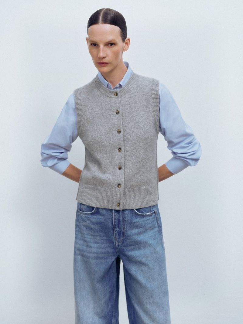 A woman in a grey knit vest and blue shirt poses in front of a white background, looking directly at the camera in a fashion shot.