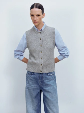 A woman in a grey knit vest and blue shirt poses in front of a white background, looking directly at the camera in a fashion shot.