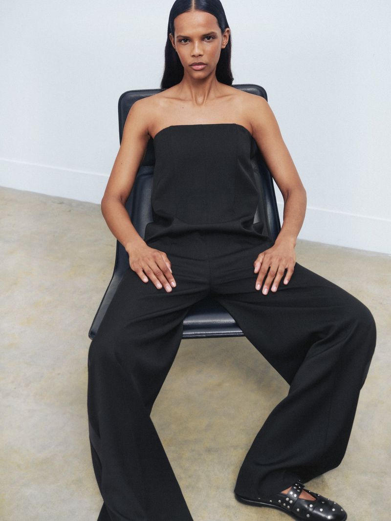 Adult woman in a black tube top and trousers sitting on a black chair in front of a white wall in a minimalist studio setting.