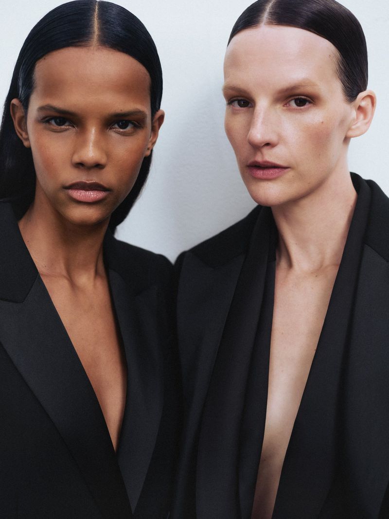 Two female models in black blazers pose in a portrait. One woman has dark skin and dark hair, the other has light skin and light hair.