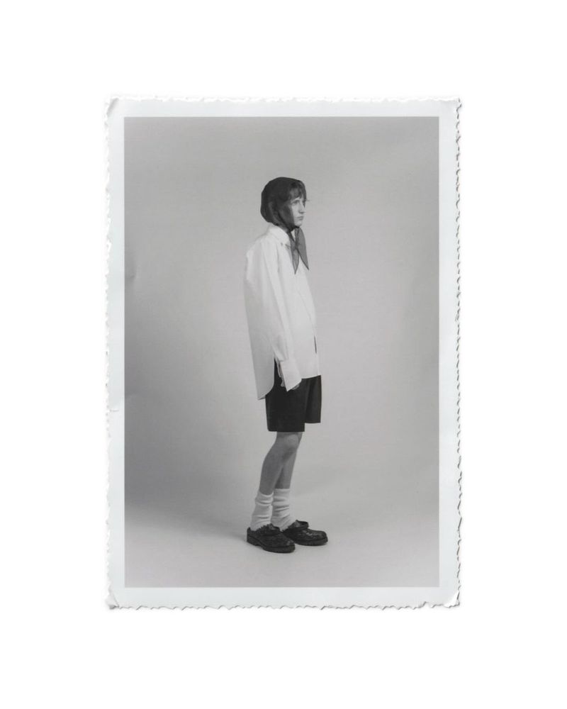 Monochrome photograph of a young adult woman in an oversized white shirt, black shorts, socks, and black shoes, posing in a studio.