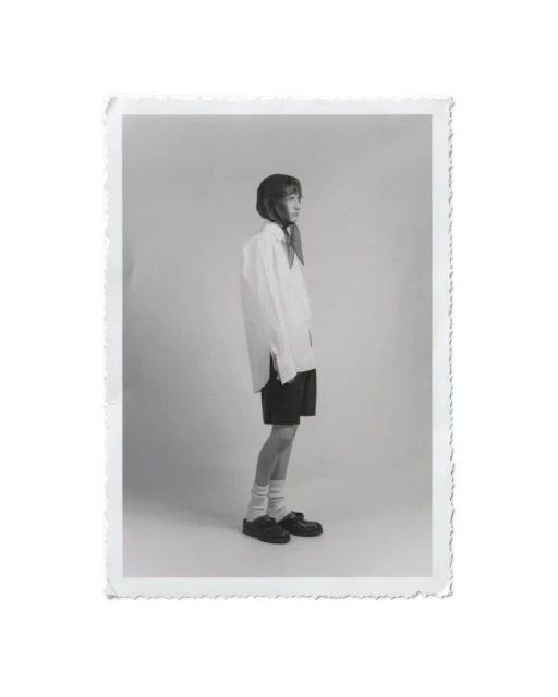 Monochrome photograph of a young adult woman in an oversized white shirt, black shorts, socks, and black shoes, posing in a studio.