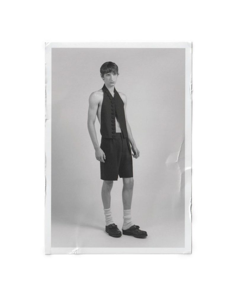 Monochrome portrait of a model standing in a black halter-neck vest and tailored shorts against a studio backdrop.