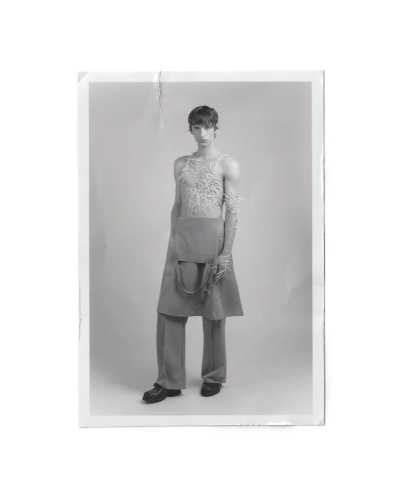 Studio shot of androgynous person wearing conceptual grey and white top, draped skirt, trousers and shoes, against a white background.