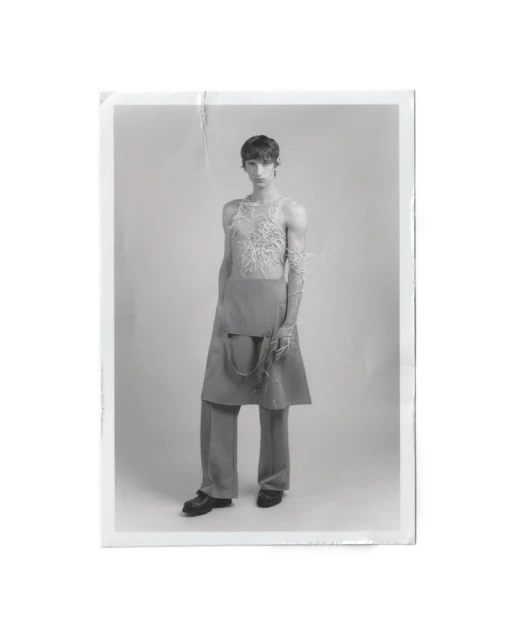 Studio shot of androgynous person wearing conceptual grey and white top, draped skirt, trousers and shoes, against a white background.