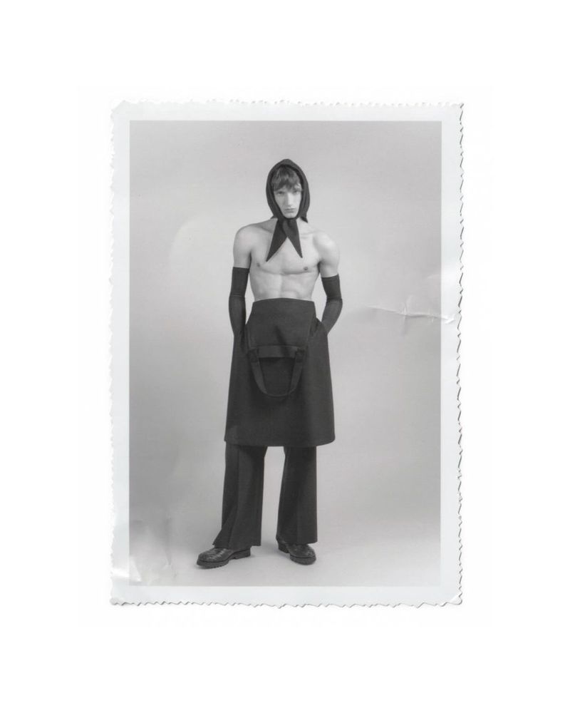 Monochrome studio portrait of a slender adult male fashion model posing shirtless with a headscarf, apron, and gloves.
