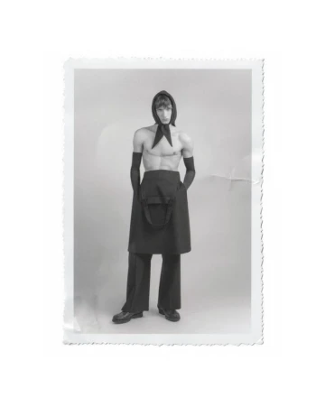 Monochrome studio portrait of a slender adult male fashion model posing shirtless with a headscarf, apron, and gloves.