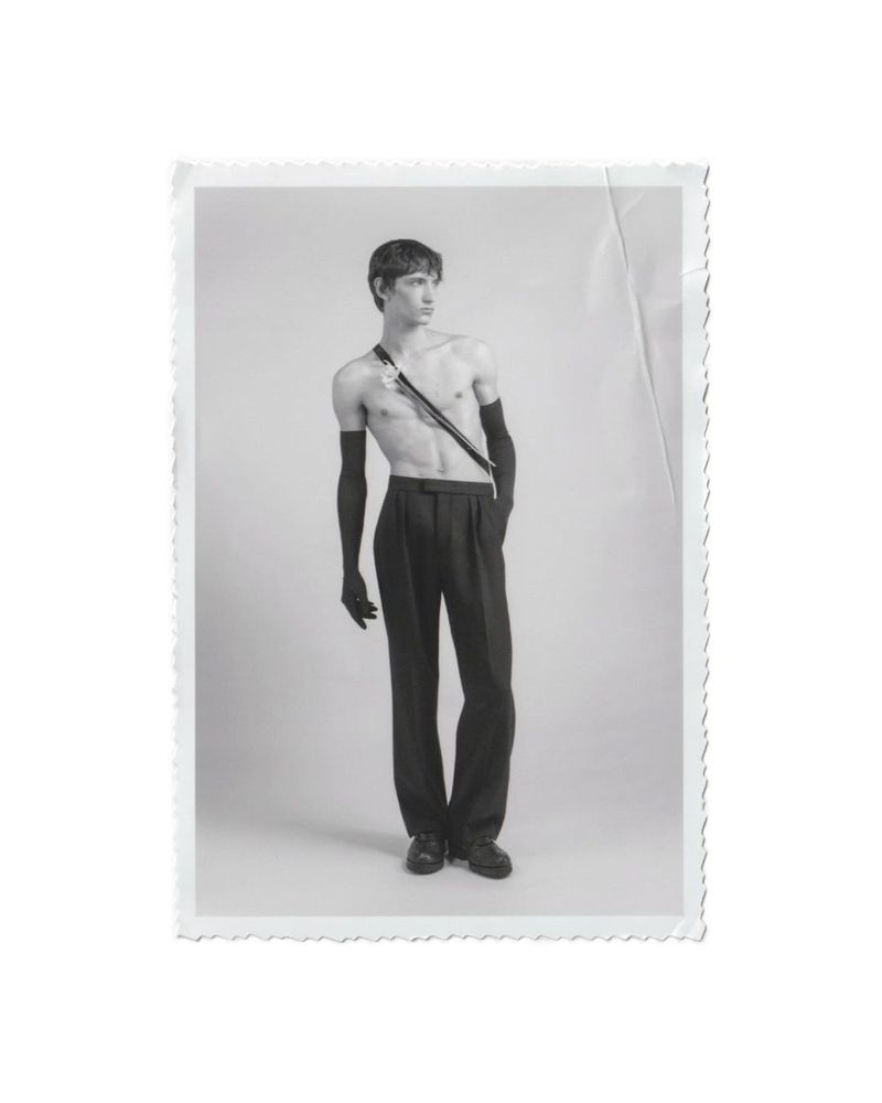 Monochrome portrait of a shirtless young man with long black gloves and dress pants, gazing off to the side in a photo studio.