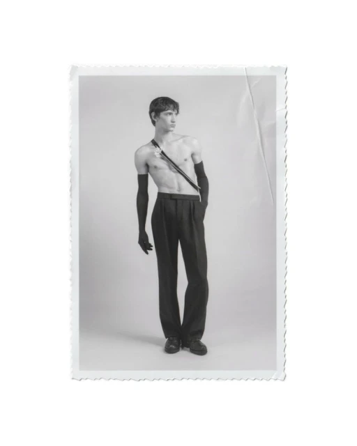 Monochrome portrait of a shirtless young man with long black gloves and dress pants, gazing off to the side in a photo studio.