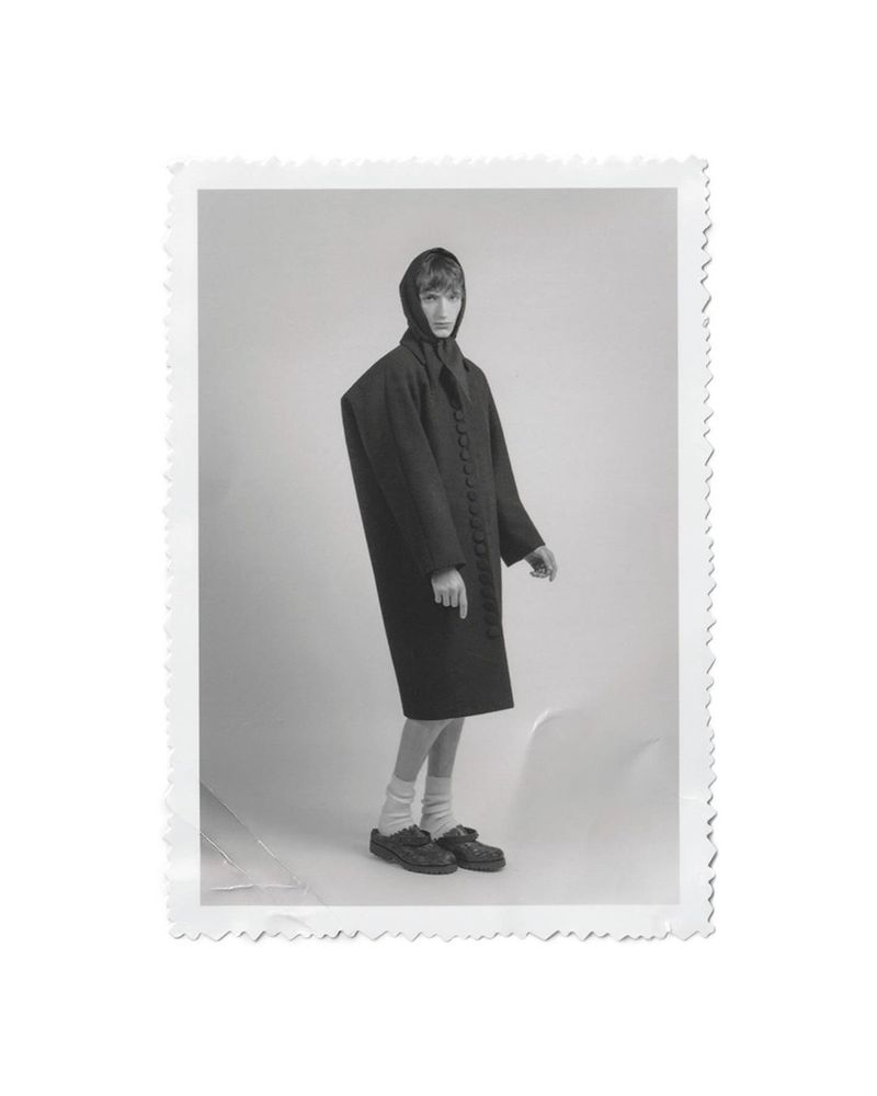 Model Tristan Gibert standing in an oversized dark buttoned coat and headpiece on a light studio background with a deckled edge.
