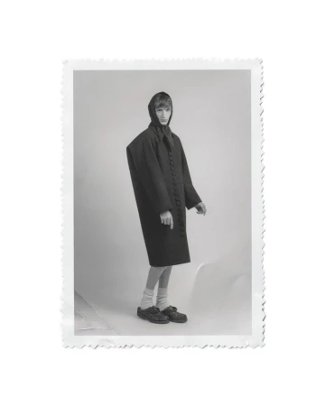Model Tristan Gibert standing in an oversized dark buttoned coat and headpiece on a light studio background with a deckled edge.