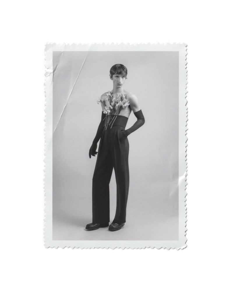 Black-and-white studio portrait of a young adult male wearing a corset, black gloves and trousers, adorned with flowers, posing elegantly.