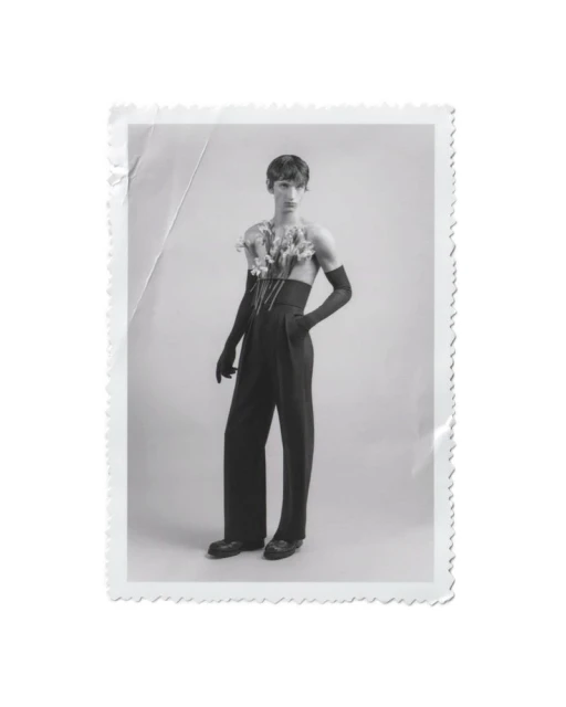 Black-and-white studio portrait of a young adult male wearing a corset, black gloves and trousers, adorned with flowers, posing elegantly.